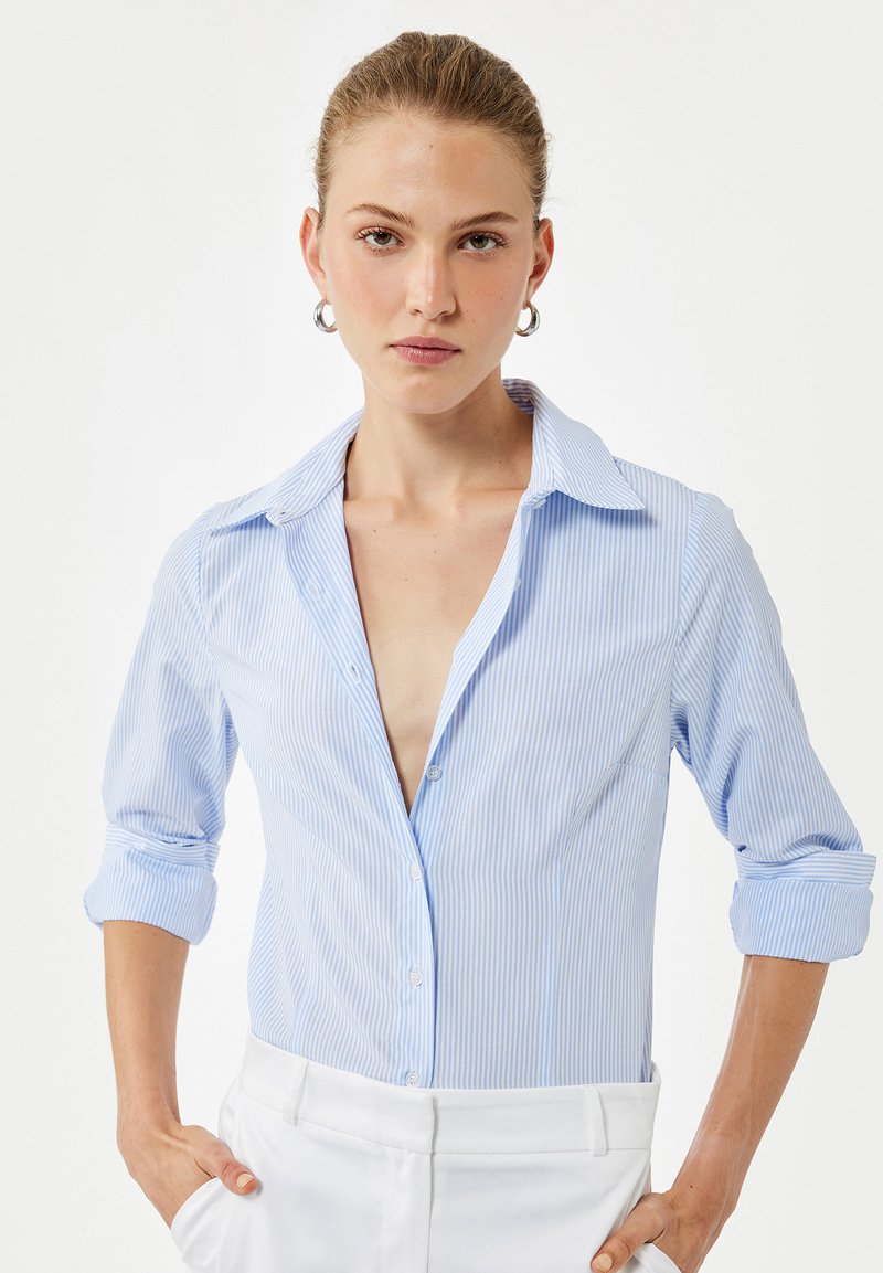 Camisa de botones a rayas azul claro con un escote en V profundo, mangas enrolladas y pantalones blancos. Tejido suave con una textura lisa.