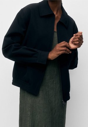 Personne portant une veste noire zippée par-dessus une robe vert foncé texturée, ajustant un poignet avec l'autre main sur un fond uni.