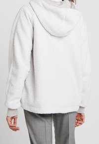 Bílá plyšová fleecová bunda s kapucí, která má texturovaný povrch a pohodlný střih. Bunda má žebrované manžety a zaoblený spodní lem.