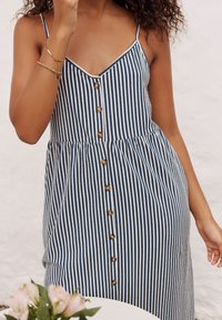 Robe à rayures bleues et blanches avec bretelles fines, ouverture boutonnée sur le devant et taille froncée. Tissu léger avec une silhouette fluide.