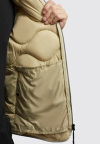 Beige pufferjack interior met golvenpatroon vulling en ritssluiting. Bevat een binnenzak en een soepele, gewatteerde textuur.