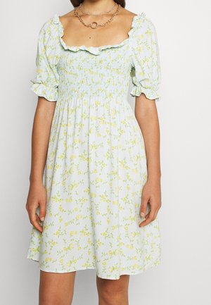 Frau trägt ein hellblaues Kleid mit gelbem Blumenmuster, Puffärmeln, gerafftem Oberteil und gerafftem Ausschnitt, kombiniert mit mehrlagigen Goldketten.