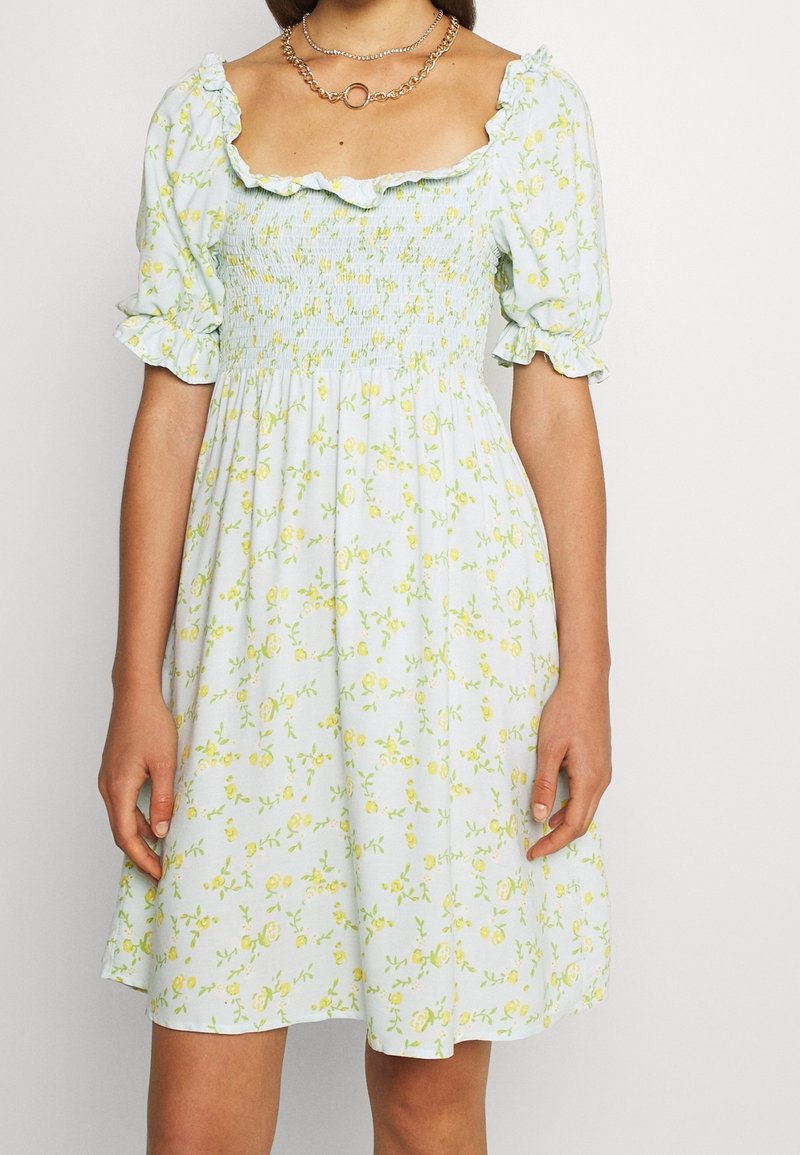 Femme portant une robe bleu clair à imprimé floral jaune, manches bouffantes, corsage froncé et encolure à volants, accessoirisée avec des chaînes en or superposées.
