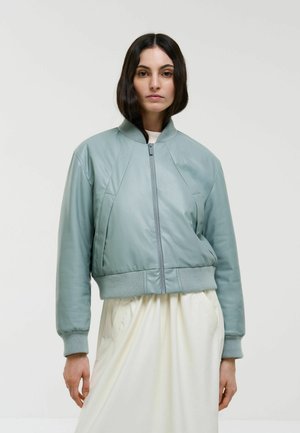 Giacca in pelle bomber di colore verde menta, con polsini e orlo a costine, caratterizzata da chiusura con zip e design a pannelli. Indossata sopra un vestito bianco.
