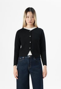 Jeune femme aux cheveux blonds raides portant un cardigan noir boutonné sur un haut blanc, associé à un jean bleu foncé taille haute, le tout sur fond blanc.