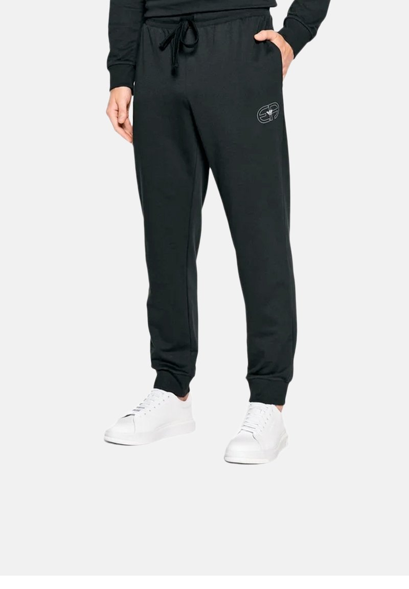 Pantalons de survêtement noirs avec une taille élastique et un cordon de serrage, arborant un logo subtil sur la cuisse, associés à des baskets blanches.