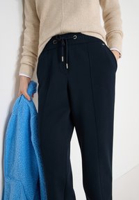 Personne portant un pull côtelé beige et un pantalon noir à cordon, tenant une veste bleue texturée dans la main gauche, main droite dans la poche.