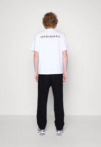 Camiseta blanca de algodón con texto rojo "ABERCROMBIE NEW YORK" en la parte posterior, combinada con pantalones negros de corte holgado y zapatillas.