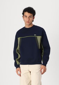 Sweatshirt bleu marine avec des accents verts, présentant un design à panneaux texturés, un col rond et des poignets côtelés. Logo Lacoste sur la poitrine.