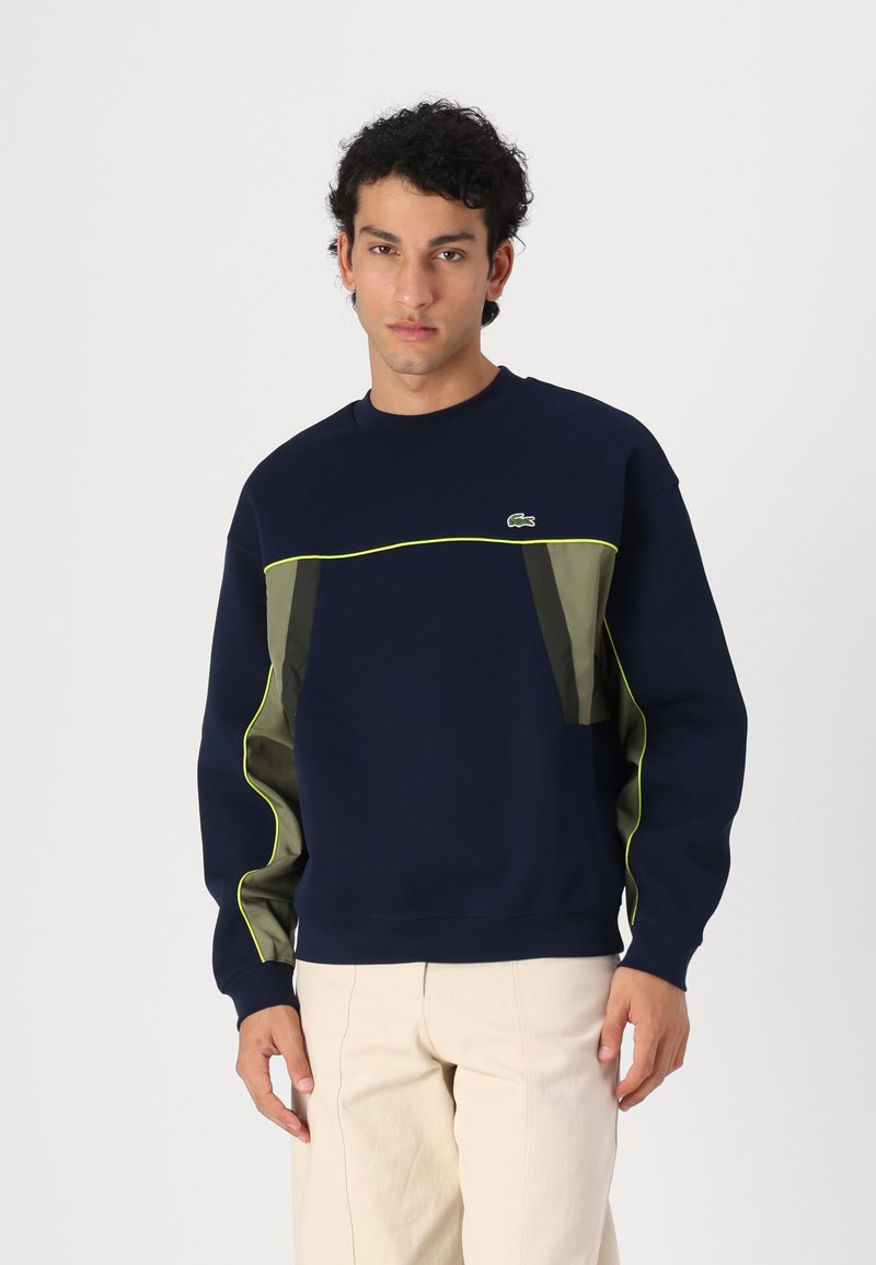Sweatshirt bleu marine avec des accents verts, présentant un design à panneaux texturés, un col rond et des poignets côtelés. Logo Lacoste sur la poitrine.