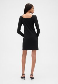 Svart langermet kjole med firkantet ryggutringning, tettsittende design og glatt tekstur. Kombinert med svarte sandaler med reimer og høye hæler.