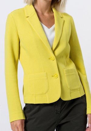 Kvinde iført en tekstureret gul blazer med to knapper foran, lommer med lapper og en hvid v-hals skjorte under.