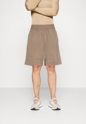 Person iført beige langærmet top, løse taupe knælange shorts og lyse beige sneakers, der står mod en ensfarvet hvid baggrund.