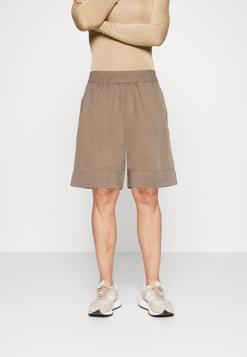 Personne portant un haut à manches longues beige, un short ample couleur taupe à hauteur des genoux, et des baskets beige clair, debout sur un fond blanc uni.