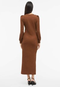 Ruskea neulottu mekko, jossa pitkät hihat, vartalonmyötäinen siluetti ja takana halkio. Sileä pinta, jossa hienovarainen pystysuuntainen kuviointi. Mustat, remmillä varustetut korkokengät.