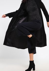 Woman wearing black sparkly top, wide-leg pants, ankle boots, and long black coat, lifting one leg mid-step against white background.