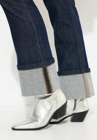 Botines metálicos plateados con punta afilada y tacón grueso, combinados con jeans de mezclilla oscuros que tienen un dobladillo gris y costuras naranjas.