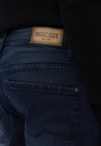 Tummansiniset farkut, joissa on nahkalappu vyötäröllä, jossa lukee "INDICODE EST. 1993", ja hienovaraisia ompeluyksityiskohtia.
