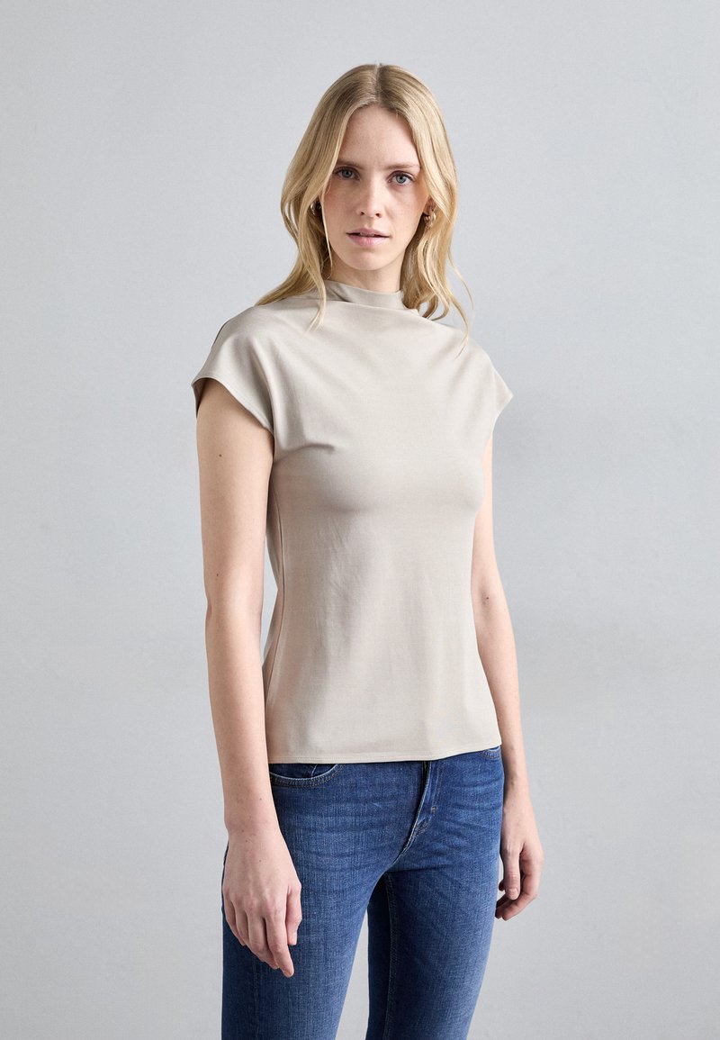 Top beige a maniche corte con collo alto, realizzato in tessuto morbido. Presenta una vestibilità aderente con una texture liscia, abbinato a jeans blu.
