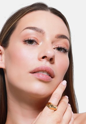 Goldring am Finger, mit einem glatten, geschwungenen Design. Nahaufnahme eines Gesichts mit neutralem Make-up, das Lippen und Augen hervorhebt. Heller Hautton.