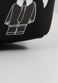 Article en tissu noir avec un graphisme blanc et gris représentant une personne simplifiée portant un costume et une cravate sur un fond clair uni.