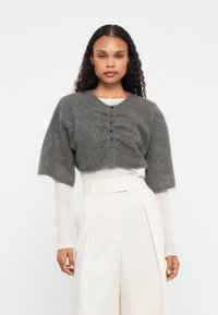 Cardigan cinza curto com mangas largas, apresentando detalhes texturizados e fecho frontal com botões. Usado sobre uma blusa de cor clara e calças.