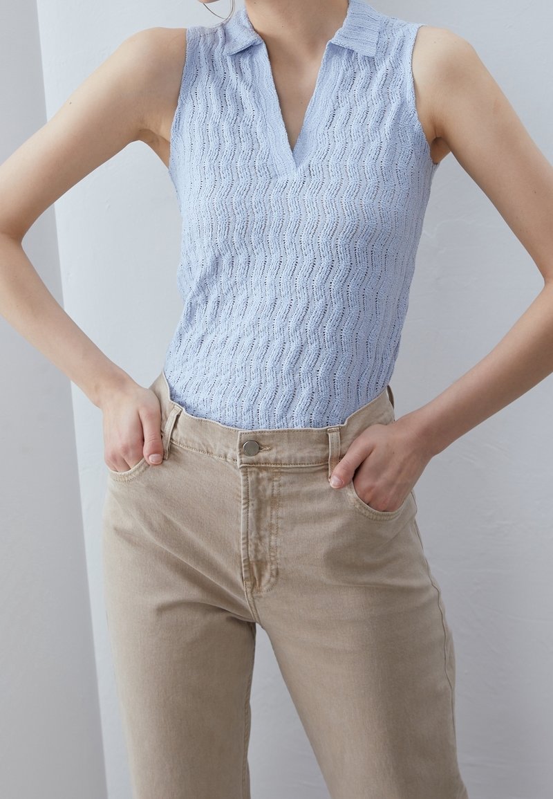 Femme portant un haut sans manches texturé bleu clair avec un col et un pantalon beige taille haute, les mains dans les poches.