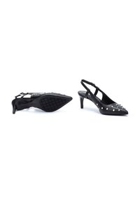 Tacones negros de piel con tira en el talón y punta afilada, adornos de tachuelas y suela texturizada. Cuenta con correa ajustable y una altura de tacón de 5 centímetros.