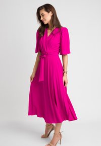 Fuksjowa sukienka w kształcie owijanej z krótkimi bufkami i związanym w pasie, z luźną spódnicą i miękkim, lekkim materiałem. Widoczne beżowe szpilki.