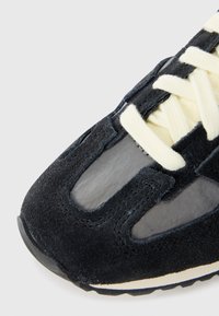 Gros plan sur l'avant d'une basket en daim noir et mesh avec des lacets de couleur crème et des coutures détaillées sur un fond clair.