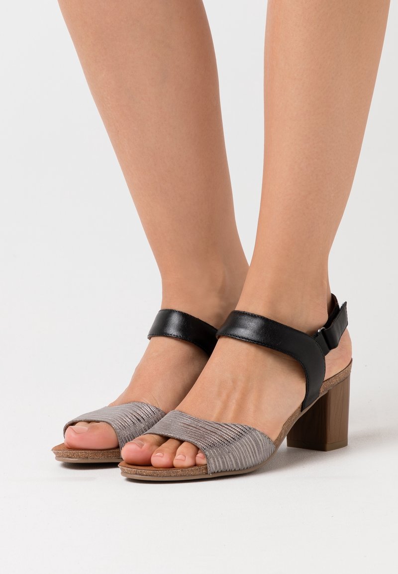 Pieds portant des sandales à bout ouvert avec des brides avant texturées grises, des brides de cheville noires et des talons blocs en bois sur fond blanc.