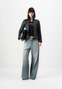 Donna che indossa un giubbotto trapuntato nero, un crop top nero, jeans a gamba larga sbiaditi, che tiene una grande pochette nera, in piedi di fronte a uno sfondo bianco semplice.