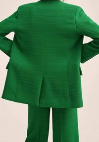 Blazer verde su misura e pantaloni coordinati realizzati in tessuto strutturato. La giacca presenta uno spacco e spalle strutturate; linee pulite e taglio aderente.
