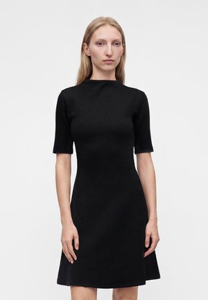 Femme aux cheveux blonds et raides portant une robe noire à manches courtes, debout devant un fond clair uni.