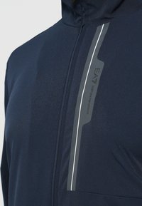 Tamsiai mėlynas vėjui atsparus švarkelis su sklandžiu paviršiumi, su sidabrine atspindinčia juostele ir EA Emporio Armani logotipu prie priekinio užtrauktuko.