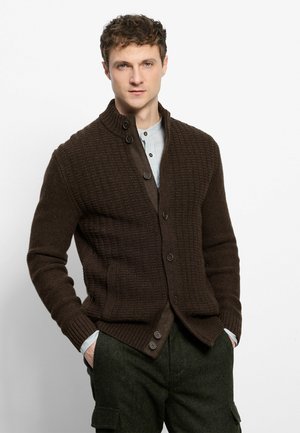 Junger Mann mit lockigem Haar, der eine dunkelbraune strukturierte, geknöpfte Strickjacke über einem hell gestreiften Hemd und dunkelgrünen Hosen trägt, Hände in den Taschen.