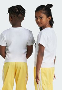 T-shirt bianchi a maniche corte indossati da due bambini, abbinati a pantaloni gialli con strisce bianche. Tessuto di cotone morbido, vestibilità rilassata.