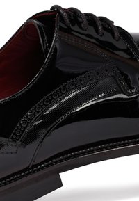 Zwarte patenten leren schoenen met decoratieve perforaties en textuurelementen, voorzien van zwarte veters en een lage hak.
