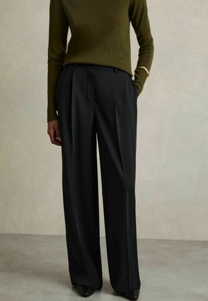 Personne portant un pantalon noir taille haute à jambes larges, un haut à manches longues vert olive, des chaussures pointues noires et un bracelet doré au poignet.
