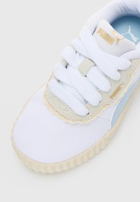 Detailný záber na biele tenisky Puma s zvlnenými béžovými semišovými prekrýtami, hrubými bielymi šnúrkami, modrým logom a ribbovanou béžovou podrážkou.