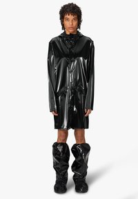 Chubasquero negro, brillante y largo, hecho de un material brillante, con capucha, bolsillos y combinado con botas de lluvia brillantes hasta la rodilla que hacen juego.