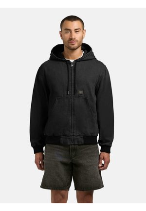 Mand iført sort hættetrøje med lynlås og mørkegrå denimshorts, stående mod en ensfarvet lys baggrund.