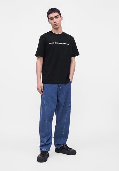 Νέος άνδρας στέκεται φορώντας μαύρο T-shirt Emporio Armani, φαρδιά μπλε τζιν και μαύρα αθλητικά παπούτσια σε απλό ανοιχτό φόντο.