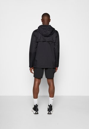 Uomo che indossa una giacca nera con cappuccio, pantaloncini neri, calzini bianchi alti e scarpe da ginnastica nere alte, in piedi di fronte a una parete bianca liscia.