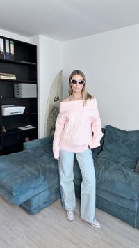 Pulover roz deschis, decupat pe umeri, din tricot cu fir prins, cu croială lejeră, asortat cu blugi cu picioare largi, de culoare albastru deschis, și adidași albi, pe un fundal de canapea teal.