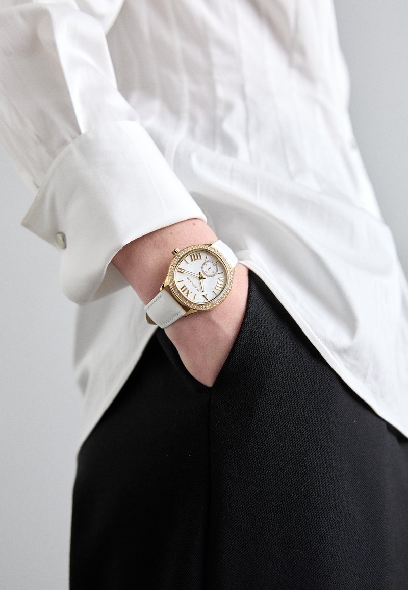 Orologio dorato con quadrante bianco e numeri romani, dotato di un cinturino in pelle bianca, posizionato su una camicia bianca e pantaloni neri.