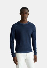 Maglione a maglia strutturata blu navy con scollatura rotonda e maniche lunghe a raglan, abbinato a jeans bianchi. Presenta polsini e orlo a coste.