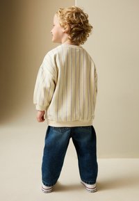 Krémová mikina s modrými svislými pruhy, volný oversized střih, doplněná volnými modrými džínami a bílými teniskami s červenými detaily.