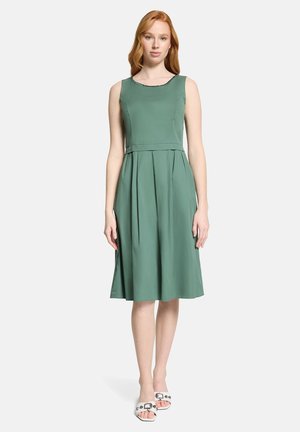 Kobieta z rudymi włosami, ubrana w zieloną, bezrękawnikową sukienkę długości do kolan oraz białe sandały na odkryte palce, stojąca na tle jasnej, jednolitej ściany.