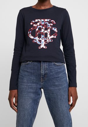 Personne portant une chemise bleu marine à manches longues avec un grand monogramme multicolore à motif floral et un jean bleu.